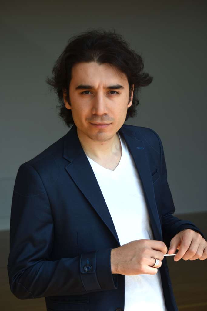 Homem posa para um retrato, usando blazer azul-marinho sobre uma camiseta branca, em frente a um fundo escuro.
