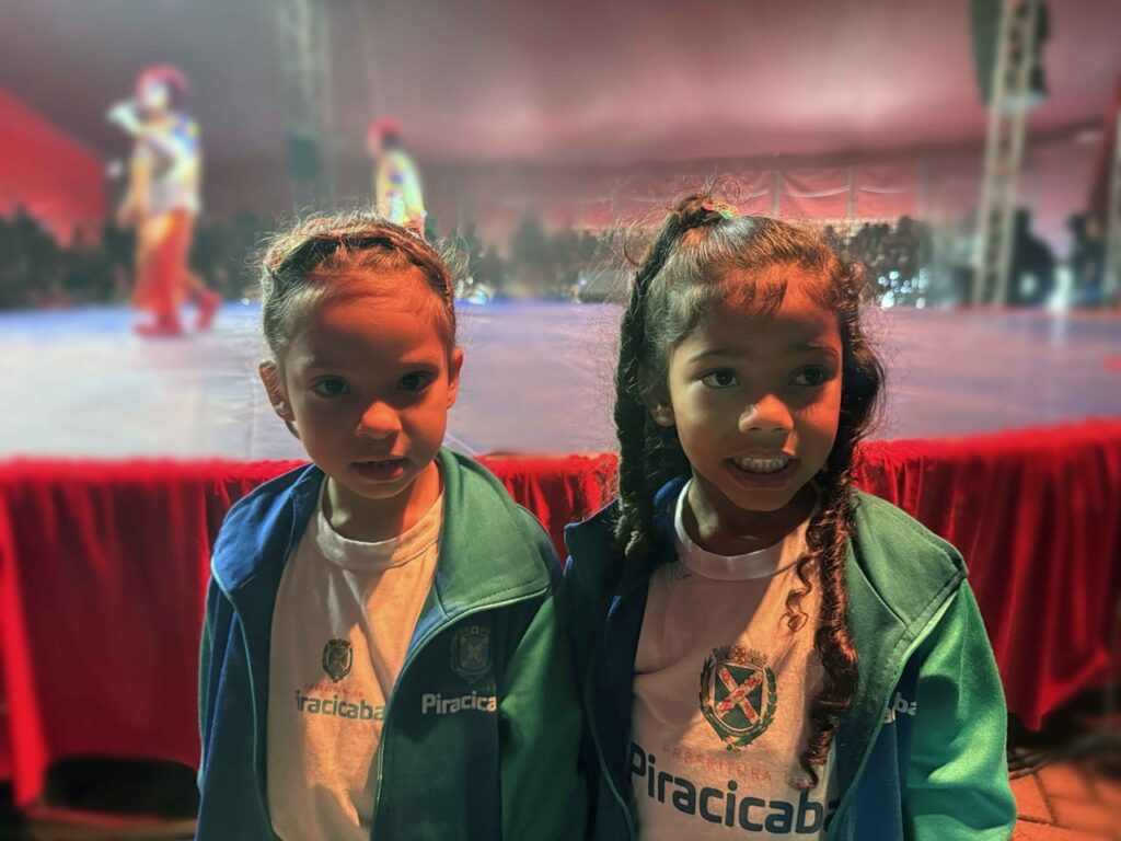 A imagem mostra duas meninas em primeiro plano, diante de um palco de circo. Elas usam jaquetas verdes e camisetas brancas com o brasão e o nome “Piracicaba”. A menina à esquerda tem o cabelo preso, enquanto a menina à direita usa duas tranças. Ao fundo, o palco está iluminado com luzes coloridas e há artistas vestidos de palhaço se apresentando, com a plateia parcialmente visível. A parte frontal do palco é decorada com um tecido vermelho.