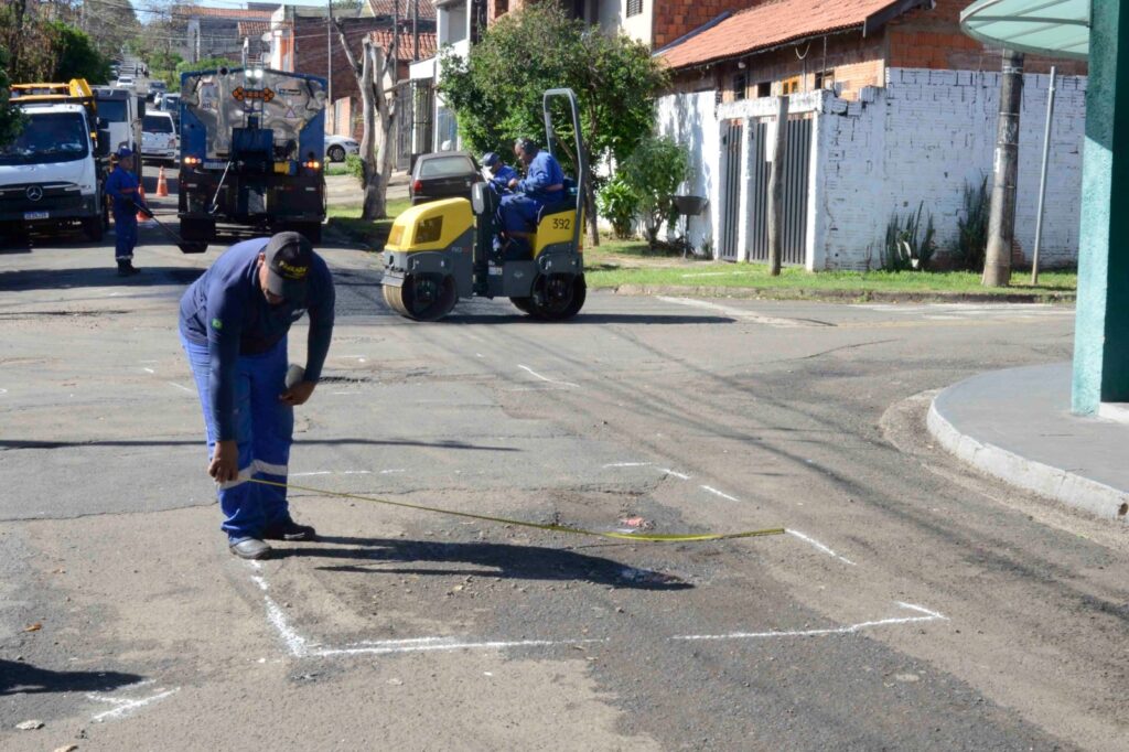 A imagem mostra trabalhadores realizando manutenção viária em um bairro residencial: no primeiro plano, um deles mede e marca com giz branco um retângulo no asfalto, enquanto ao fundo aparecem veículos como um rolo compressor e caminhões, além de outros operários envolvidos no reparo da rua.