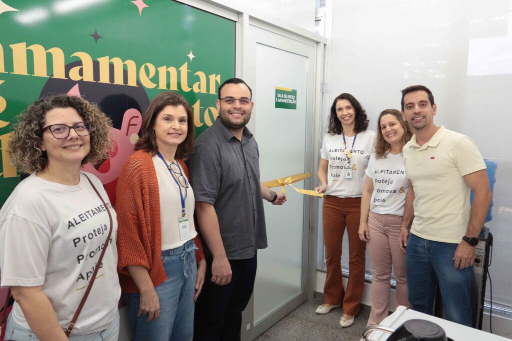 A imagem mostra uma foto de grupo em um ambiente interno, provavelmente durante um evento ou encontro profissional. Várias pessoas estão reunidas e sorrindo para a câmera, usando crachás e camisetas com a palavra “ALEITAMENTO” e referências a “Promova”. Ao fundo, há um grande painel verde com letras amarelas, ilustrações coloridas e a indicação “Sala de Apoio à Amamentação”. A foto está enquadrada de forma bastante aproximada e aparece girada de lado.
