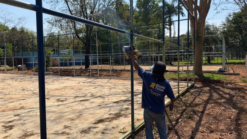 Trabalhador realiza a pintura da estrutura metálica de uma quadra esportiva em área que passa por reformas e melhorias.