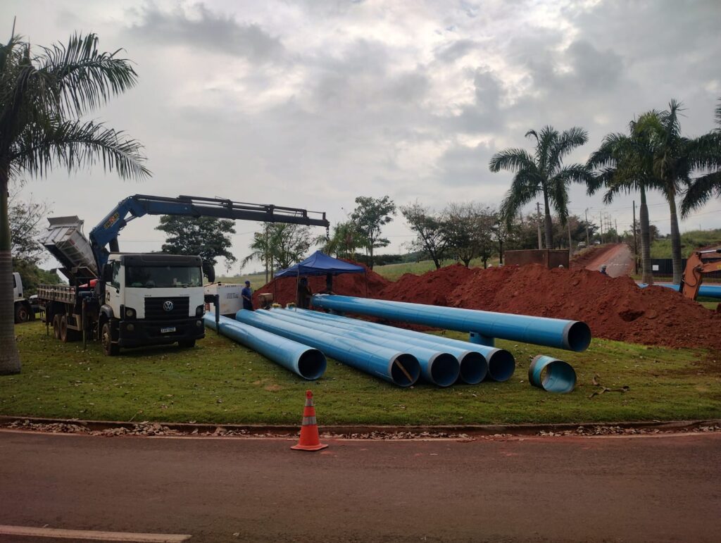 A cena captura um canteiro de obras com materiais de construção e equipamentos. Em primeiro plano, há uma estrada de asfalto em tons de marrom avermelhado. Ao lado da estrada, uma área gramada verde contém vários tubos grandes e azuis dispostos horizontalmente. Há também um cone de trânsito laranja próximo aos tubos. O foco principal da imagem está em um conjunto de tubos azuis de grande diâmetro deitados na grama. Eles parecem estar sendo preparados para instalação. Um dos tubos, mais à direita, está ligeiramente separado dos outros e parece inclinado. Um caminhão branco com uma carroceria aberta e um guindaste azul acoplado está posicionado à esquerda da imagem. O guindaste está levantando ou descarregando material da carroceria do caminhão. Uma pessoa com camisa azul e calça escura está perto do caminhão, observando o trabalho. Há também uma pequena tenda azul montada em segundo plano, provavelmente para fornecer abrigo. O fundo mostra uma paisagem com montes de terra avermelhada, que parecem ter sido escavados. Há palmeiras e outras árvores dispersas pela área, com folhagens verdes e algumas com aparência mais seca. O céu está nublado, com nuvens de cor cinza e branca que sugerem um dia potencialmente chuvoso ou com pouca luz solar. Ao fundo, uma estrada sinuosa se afasta para a direita, e uma máquina de construção de cor laranja pode ser vista. A composição da imagem é horizontal, com os tubos liderando o olhar do espectador através da cena. As cores predominantes são o azul dos tubos, o verde da grama, o vermelho da terra e o branco do caminhão, contrastando com o céu nublado. A imagem transmite uma sensação de trabalho em andamento e de infraestrutura sendo construída.