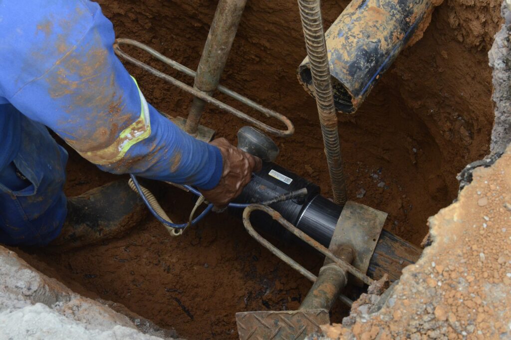 A imagem mostra um trabalhador realizando a instalação de uma tubulação subterrânea dentro de uma vala escavada. O profissional utiliza equipamentos e ferramentas para conectar a nova rede de água por meio do método não destrutivo de furo direcional, em uma obra de infraestrutura de saneamento.