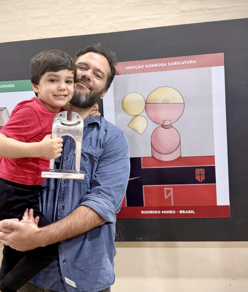 Uma pessoa segura uma criança no colo enquanto exibe um troféu em frente a um painel de exposição. Ao fundo, há um cartaz de uma caricatura com a inscrição “Menção Honrosa Caricatura”.