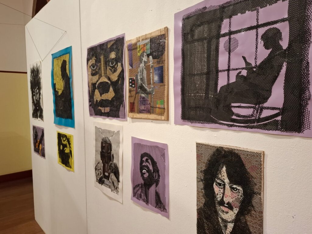 Galeria de arte com uma grande obra têxtil colorida retratando um rosto, cercada por outras ilustrações e trabalhos artísticos em exibição.