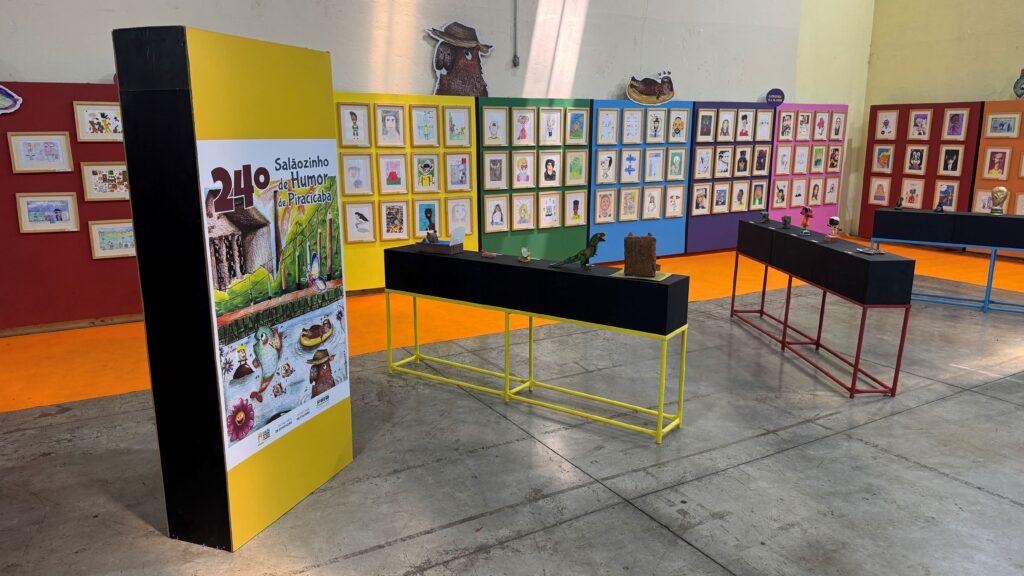 A imagem mostra uma exposição de arte infantil em um amplo espaço interno. Nas paredes coloridas estão expostos diversos desenhos emoldurados, organizados em painéis vermelhos, amarelos, verdes, azuis e roxos.