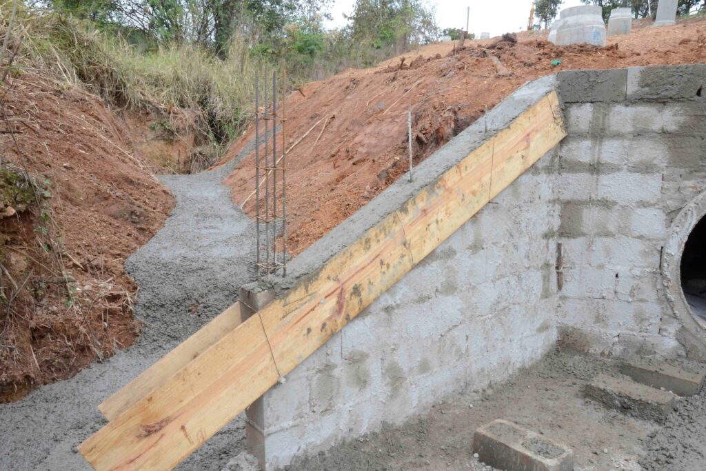 A parede de concreto está em construção com vergalhões e formas de madeira. O tubo circular indica parte de um sistema de drenagem ou bueiro. O cenário mostra uma etapa inicial da engenharia civil voltada para infraestrutura.