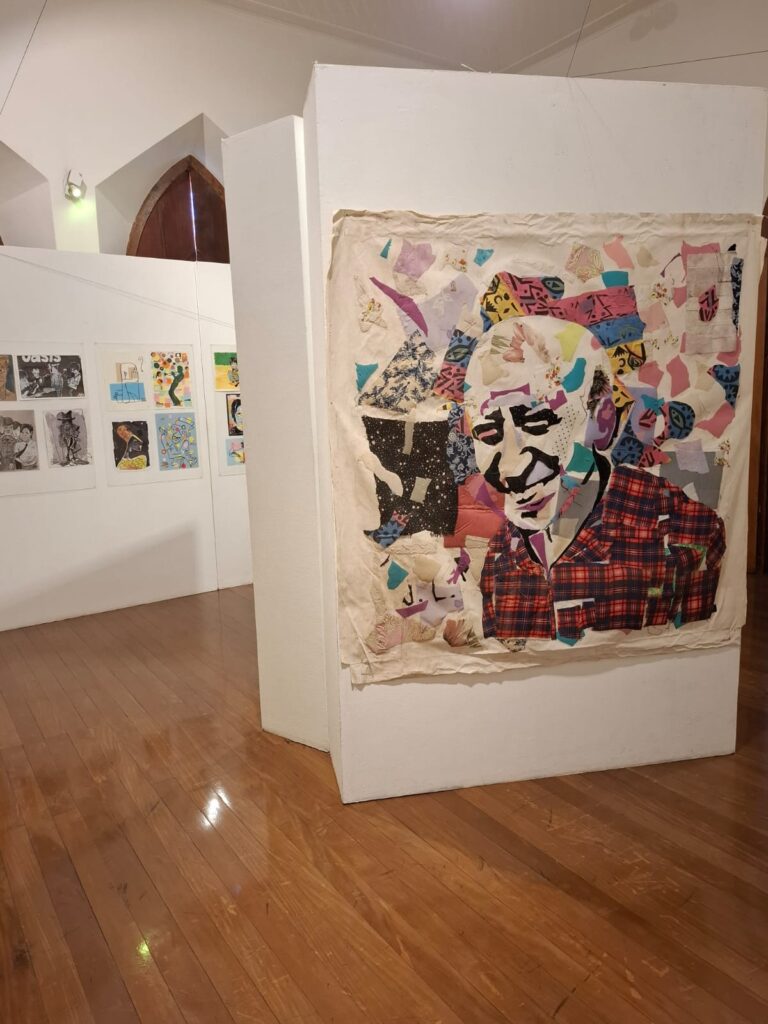 Galeria de arte com uma grande obra têxtil colorida retratando um rosto, cercada por outras ilustrações e trabalhos artísticos em exibição.