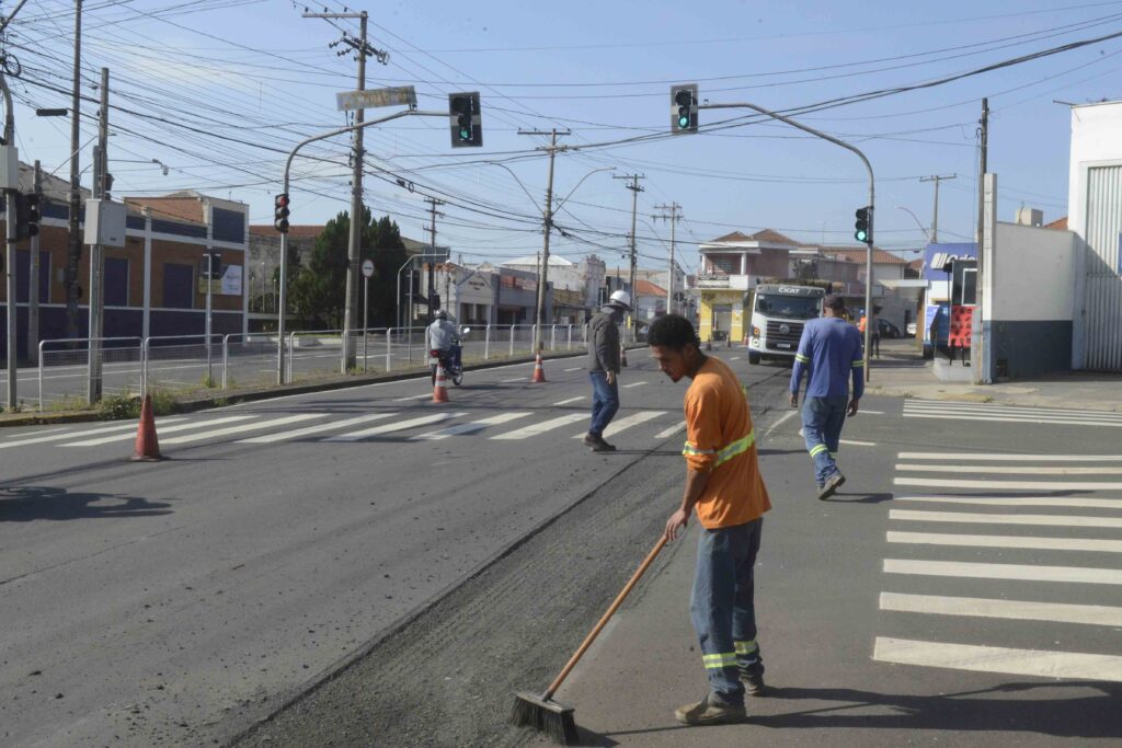 A imagem mostra trabalhadores realizando obras de recapeamento em uma avenida urbana.