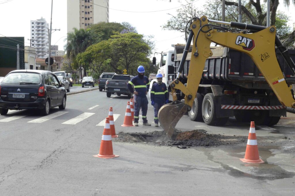 A imagem mostra uma obra viária em uma avenida urbana. Dois trabalhadores com equipamentos de segurança acompanham uma retroescavadeira que realiza escavações no asfalto, enquanto cones de sinalização isolam a área. Ao redor, há veículos em circulação, árvores e edifícios ao fundo.