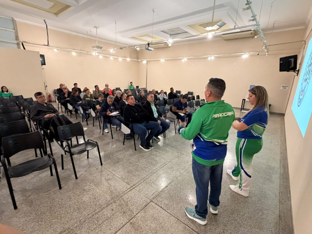 A imagem mostra uma apresentação em uma sala de eventos, onde duas pessoas — vestindo uniformes esportivos nas cores verde, azul e branco com o nome “Piracicaba” — falam diante de um grupo de participantes sentados.