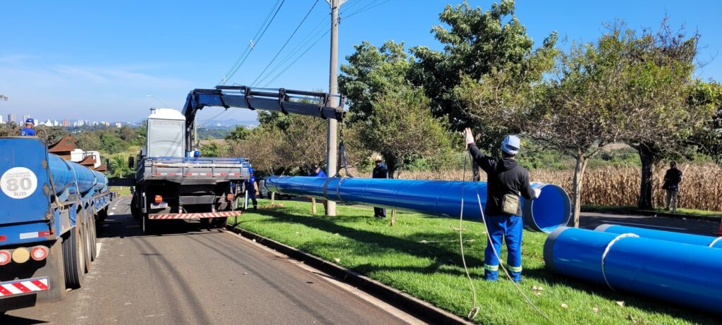 A cena externa, sob um céu azul claro, mostra uma operação de descarga de grandes tubos azuis. No lado esquerdo da imagem, vemos a traseira de um caminhão azul com um reboque, carregado com mais tubos azuis. Uma etiqueta circular branca com a inscrição "VELOCIDADE CONTROLADA 80 km" está visível na lateral do reboque. Ao lado do caminhão, um guindaste, acoplado a outro caminhão, está em operação. Este guindaste está levantando um longo tubo azul brilhante, que está sendo guiado por um trabalhador. Este trabalhador está usando um capacete azul claro, uma jaqueta preta, calças azuis com faixas refletivas amarelas nos tornozelos e luvas brancas. Ele segura uma corda de orientação. Mais à direita, vários outros tubos azuis grandes estão dispostos na grama verde, prontos para serem instalados. Há mais trabalhadores visíveis em segundo plano, perto dos caminhões e tubos, e um homem em pé ao lado de uma plantação de milho seca em segundo plano à direita. A composição da imagem é dominada pela linha horizontal dos tubos e pela linha diagonal do braço do guindaste, criando um senso de profundidade e ação. As cores predominantes são o azul vibrante dos tubos e do caminhão, o verde da grama, o marrom da vegetação ao fundo e o azul claro do céu. A luz do sol é forte, criando sombras definidas.