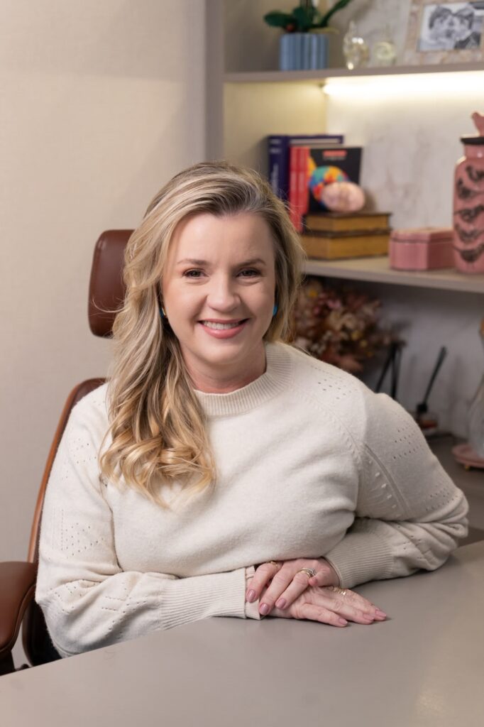 A imagem mostra uma mulher sorrindo, sentada diante de uma mesa. Ela tem cabelos loiros longos e ondulados, usa brincos azuis e um suéter claro de tricô. Suas mãos estão apoiadas sobre a mesa, e ela usa anéis e esmalte rosa-claro. Ao fundo, há prateleiras com livros, caixas decorativas, vasos, plantas e outros objetos. A iluminação é quente e o ambiente transmite uma sensação acolhedora e profissional.