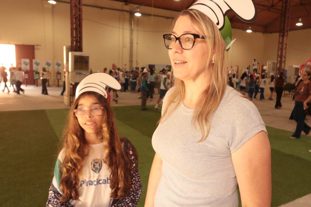 Uma criança e um adulto participam de um evento educativo em um pavilhão, usando acessórios temáticos dos Smurfs enquanto observam as atividades ao redor.