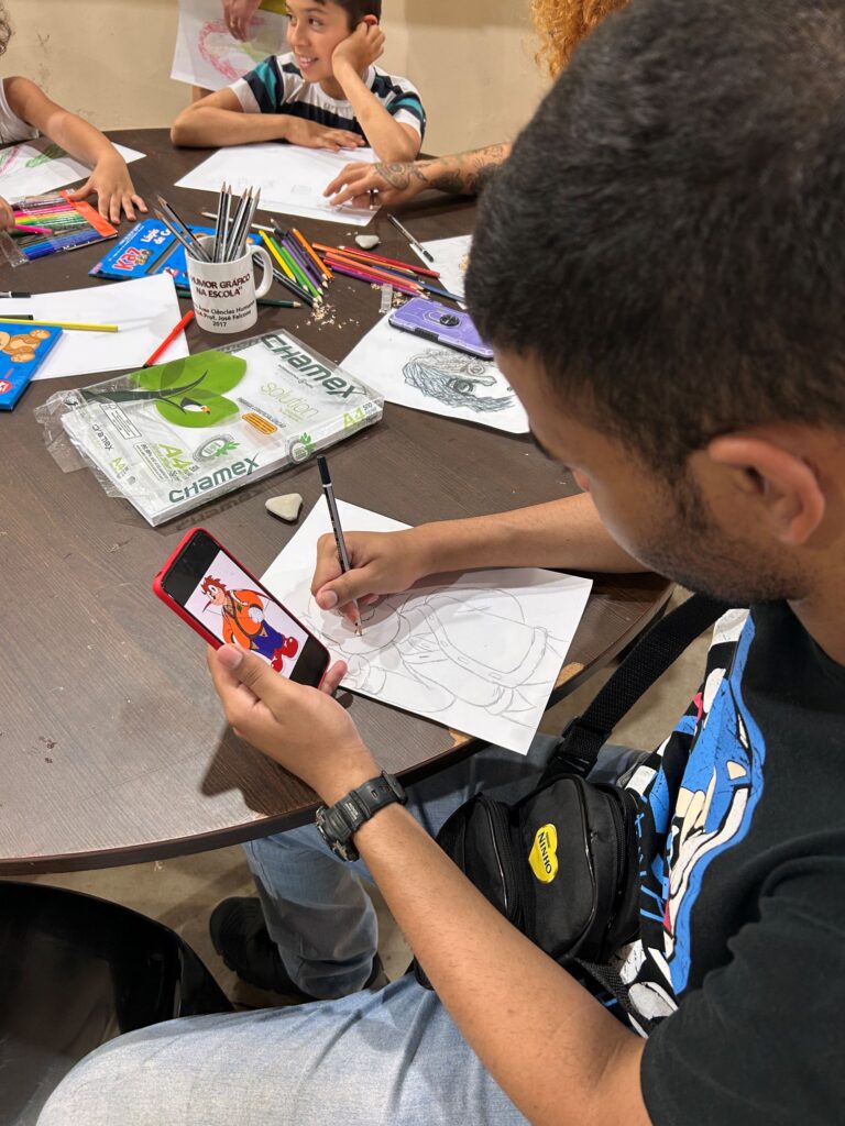Na imagem, um grupo de pessoas participa de uma atividade artística em torno de uma mesa, onde estão espalhados papéis, lápis de cor e materiais de desenho. Um dos participantes utiliza o celular para observar um personagem de desenho animado e reproduzi-lo em sua folha, enquanto outros, incluindo uma criança, também se dedicam a criar e colorir suas próprias ilustrações.