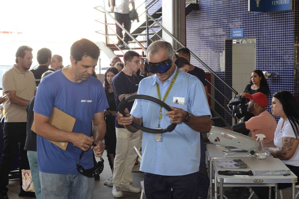 Pessoa participa de uma simulação de situações de risco no trânsito utilizando óculos que alteram a percepção visual e segurando um volante, enquanto outra pessoa acompanha a atividade. Ao redor, há participantes e um estande de serviços em um evento educativo.