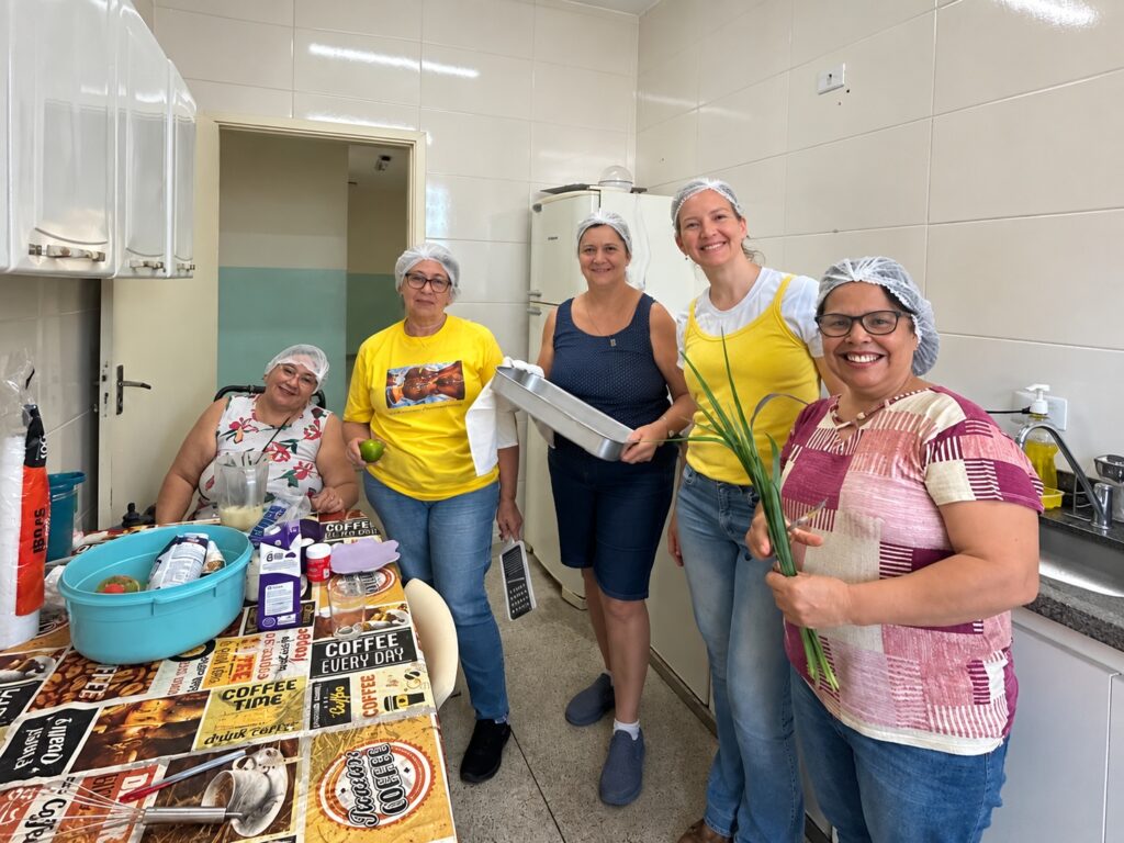 Grupo de participantes em uma cozinha comunitária durante uma oficina culinária, reunidos em torno de ingredientes e utensílios para o preparo de uma receita.