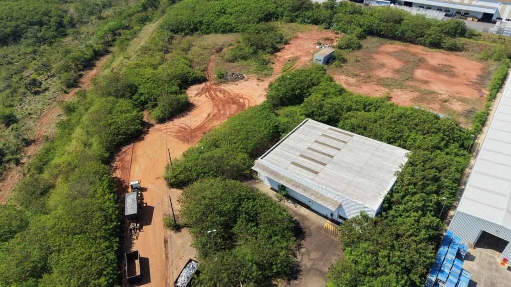 Área industrial com galpão, terreno aberto e acesso para caminhões em meio a vegetação.