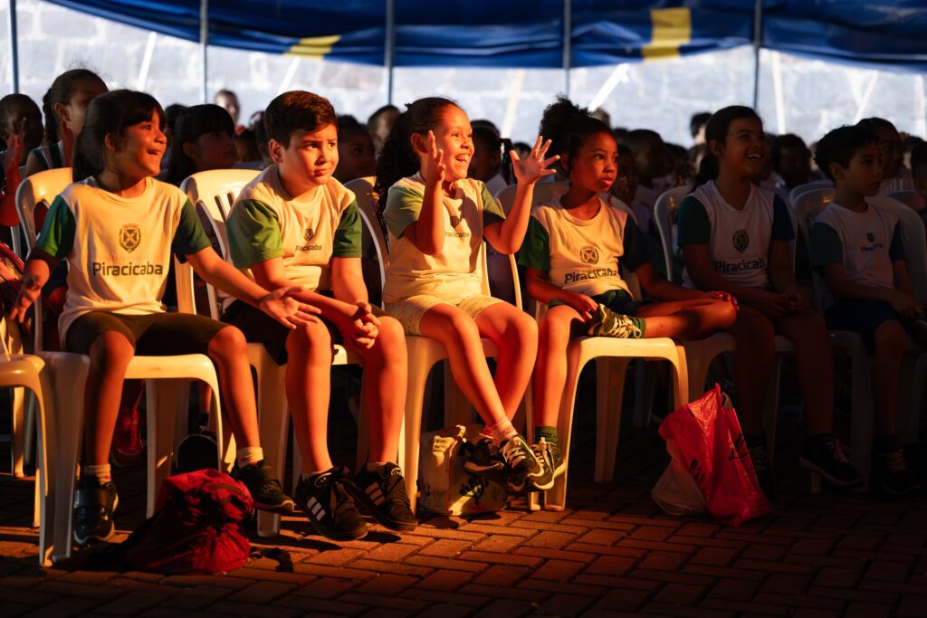 A imagem mostra um grupo de crianças de Piracicaba sentadas em cadeiras brancas dentro de uma tenda de circo, assistindo a uma apresentação. Elas vestem uniformes escolares com o nome da cidade e demonstram diferentes reações — curiosidade, surpresa e alegria — enquanto observam o espetáculo. A luz quente do ambiente destaca seus rostos e cria uma atmosfera acolhedora e divertida, típica de uma atividade cultural escolar.