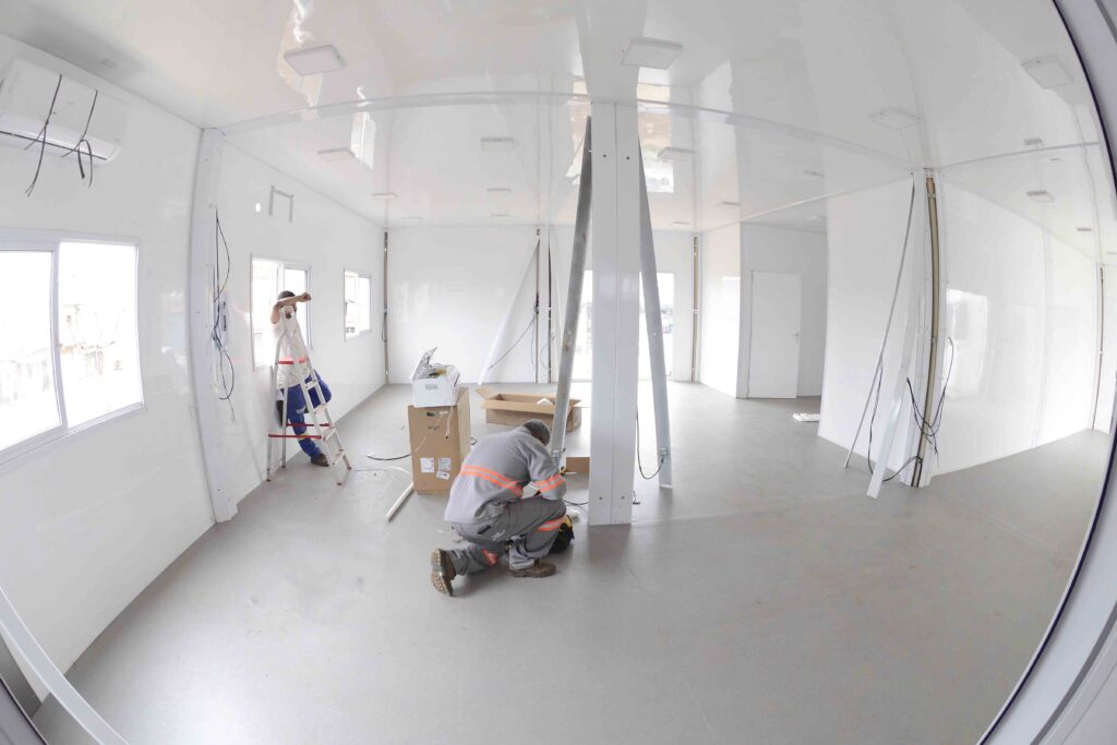 A imagem mostra dois trabalhadores realizando a instalação em um espaço modular branco. A cena se passa dentro de uma sala com paredes e teto brancos, onde a iluminação é refletida pela superfície brilhante do teto. O chão é de cor cinza claro e parece ser liso. Há janelas com molduras brancas em uma das paredes, permitindo a entrada de luz natural e mostrando o exterior com alguma vegetação e construções ao fundo. Duas pessoas estão trabalhando no local. Na lateral esquerda da imagem, um homem com uniforme branco e calças azuis está em cima de uma escada vermelha, perto de uma janela, aparentemente ajustando algo na parede. Ele usa luvas e óculos de proteção. Outro homem, vestindo um uniforme de trabalho cinza com detalhes em laranja neon, está agachado no centro da sala, concentrado em uma tarefa no chão. Ele usa sapatos de segurança marrons. Vários objetos estão espalhados pelo espaço, indicando um processo de instalação ou montagem. Há caixas de papelão, uma impressora branca sobre uma caixa, fios pretos pendurados em algumas partes, e peças modulares brancas ainda não totalmente fixadas. Uma porta branca fechada está visível ao fundo. A atmosfera é de trabalho em andamento, com um senso de construção e organização. A paleta de cores é dominada por branco e cinza, com os uniformes dos trabalhadores e a escada adicionando toques de cor. A perspectiva da imagem, ligeiramente distorcida, dá uma sensação de amplitude ao ambiente.
