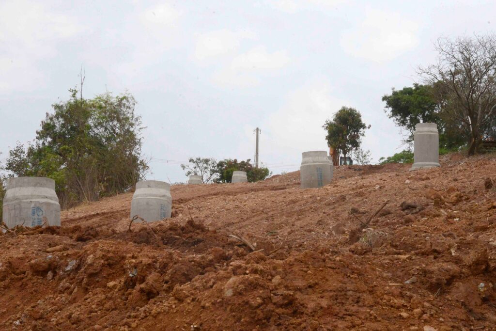 Os cilindros de concreto estão parcialmente enterrados no solo inclinado. Eles funcionam como bases de fundação para futuras obras de infraestrutura. O cenário mostra uma etapa inicial da engenharia civil em preparação do terreno.