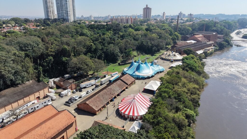 A imagem aérea mostra um complexo cultural ou de eventos às margens de um rio. No centro, há várias tendas de circo coloridas: uma azul e branca, uma vermelha e branca e outra amarela e branca. O local também possui antigos galpões de tijolos com telhados de cerâmica, veículos estacionados e áreas abertas. Ao redor, há muita vegetação e árvores. Do outro lado do rio, vê-se uma área urbana com casas, prédios residenciais altos e torres espalhadas pelo horizonte.