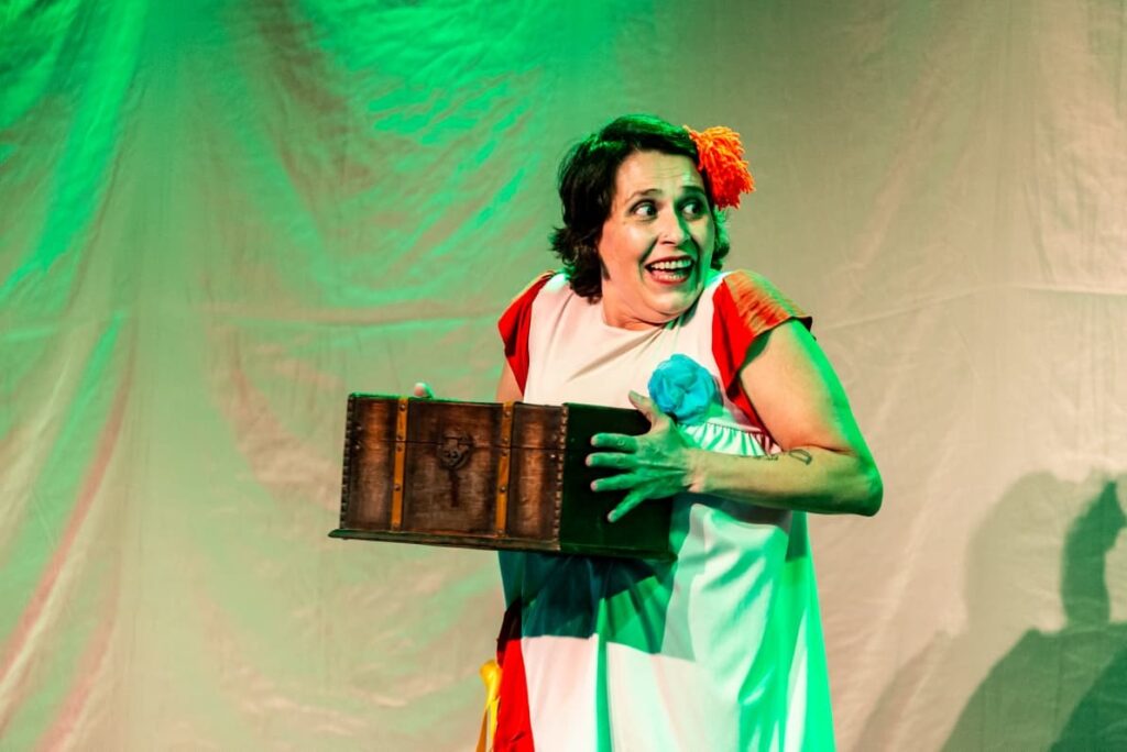 A imagem mostra uma atriz em um palco, sob iluminação verde e vermelha. Ela usa uma fantasia colorida, com vestido branco, mangas vermelhas, uma flor azul no peito e uma flor laranja presa ao cabelo. A atriz segura uma caixa de madeira escura, parecida com um baú, e olha para o lado com uma expressão surpresa e divertida. Ao fundo, há um tecido claro usado como cenário, com sombras projetadas.