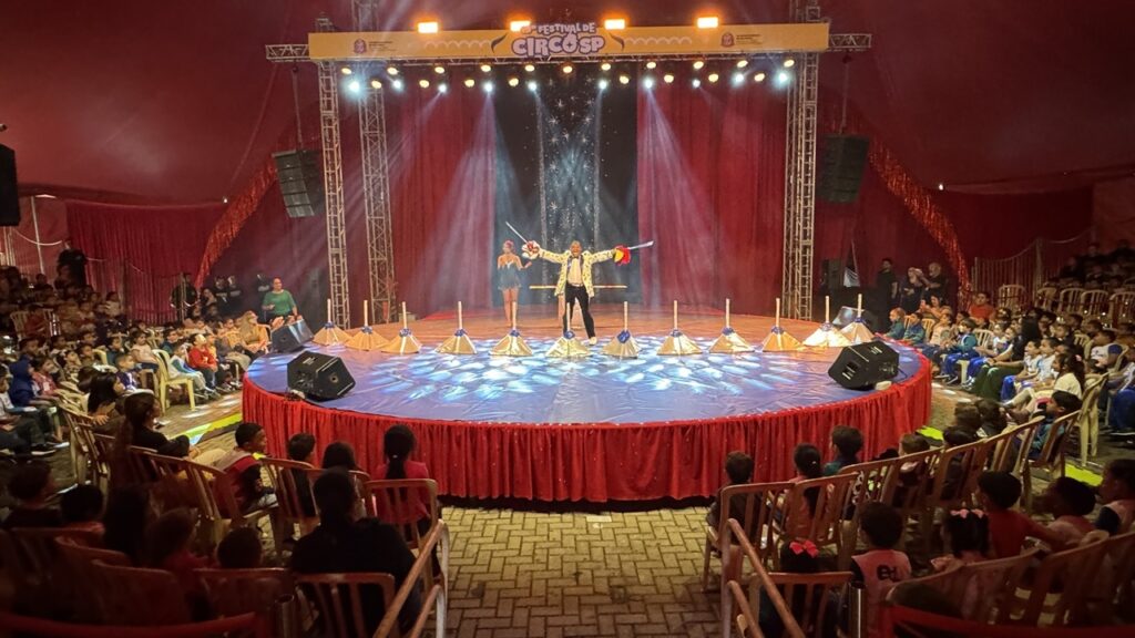 A imagem mostra um espetáculo circense acontecendo em uma grande tenda vermelha, durante o 15º Festival de Circo SP. No centro do picadeiro circular, um artista está com os braços abertos ao lado de uma mulher, diante de vários objetos coloridos usados na apresentação. O palco é iluminado por refletores azuis, brancos e amarelos, com cortinas vermelhas e um painel escuro decorado com estrelas ao fundo. O público, formado por adultos e muitas crianças, está sentado ao redor do picadeiro em cadeiras de madeira e plástico, acompanhando atentamente o espetáculo.