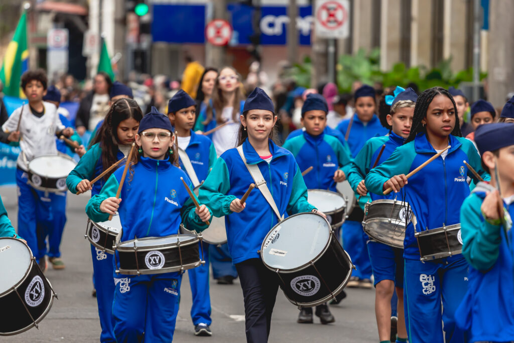 Na imagem, um grupo de crianças participa de um desfile pelas ruas da cidade, vestindo uniformes azul e turquesa com a palavra Piracicaba estampada. Elas marcham em formação tocando tambores, algumas usando bonés, criando uma apresentação musical coordenada e vibrante. Ao fundo, aparecem outros participantes, bandeiras e espectadores acompanhando o evento
