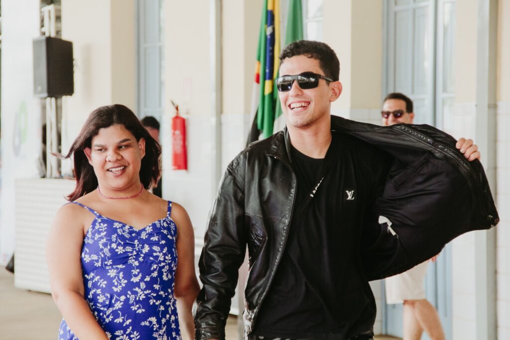 A imagem mostra duas pessoas caminhando em um ambiente interno que lembra um salão ou corredor de evento. Uma delas veste um traje azul estampado, enquanto a outra usa roupas pretas, jaqueta de couro e óculos escuros.