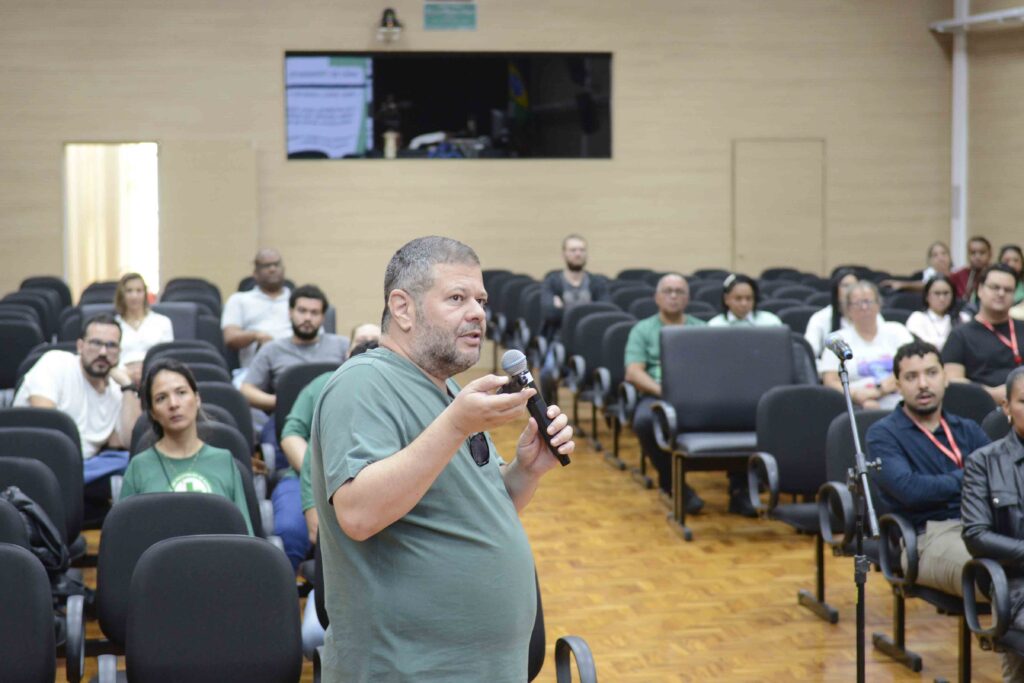 Participante faz uma pergunta ou comentário ao microfone durante uma palestra, enquanto o público acompanha a apresentação em um auditório.
