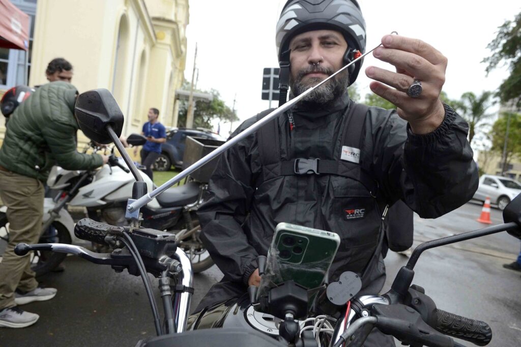 Um motociclista sentado na sua motocicleta segura e exibe a extremidade de uma antena metálica corta-cerol/corta-pipa recém-instalada no guiador da moto. Ele está equipado com casaco impermeável, capacete e tem o telemóvel num suporte no guiador.
