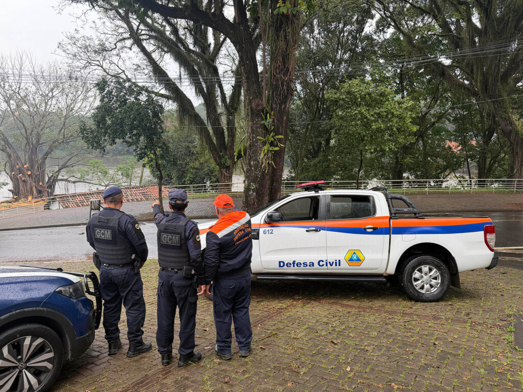 A imagem mostra dois agentes da Guarda Civil Municipal (GCM) e um integrante da Defesa Civil observando uma área próxima a um rio, em um dia nublado e chuvoso. Ao lado deles há uma caminhonete branca identificada com as cores e o logotipo da Defesa Civil. O grupo parece realizar uma inspeção ou monitoramento de segurança ambiental, reforçando a cooperação entre órgãos municipais.