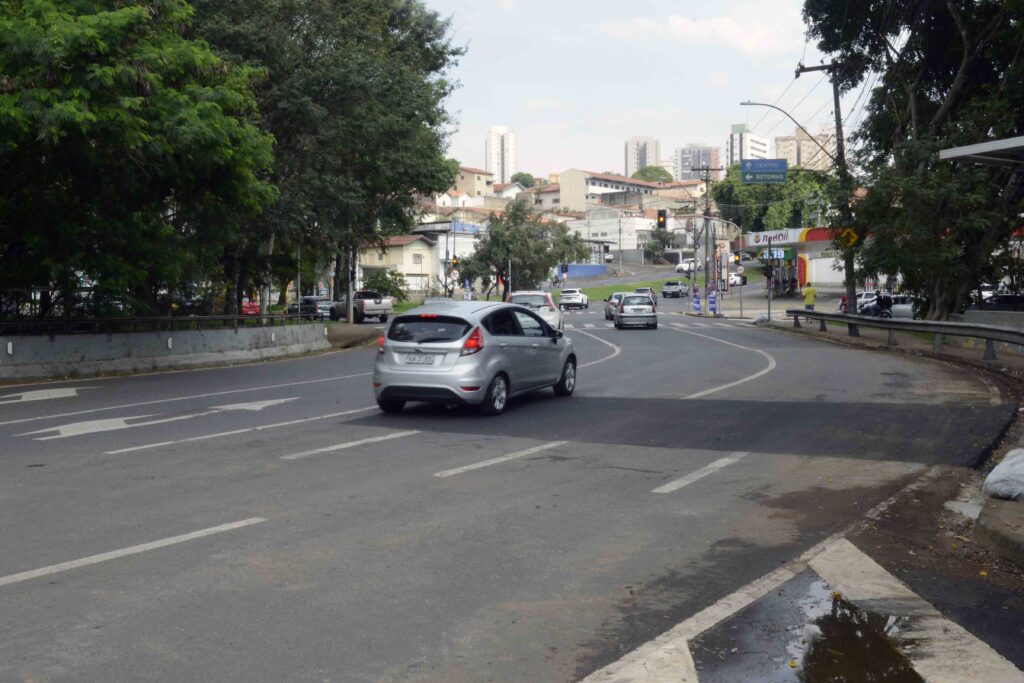 Vista de uma via asfaltada em curva com carros a circular nos dois sentidos sobre a ponte do Ribeirão Piracicamirim, destacando-se um automóvel prateado em primeiro plano e um posto de combustível ao fundo.