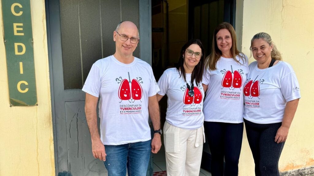 A foto mostra quatro pessoas em frente à entrada de um prédio, posando lado a lado e sorrindo. Todas usam camisetas brancas com a ilustração de dois pulmões vermelhos e a mensagem: “Desconfiar da tuberculose é o primeiro passo para começar o diagnóstico.” À esquerda, há uma placa vertical com a palavra “CEDIC”. A entrada tem uma porta escura, paredes amarelas e um pequeno piso quadriculado próximo à porta.