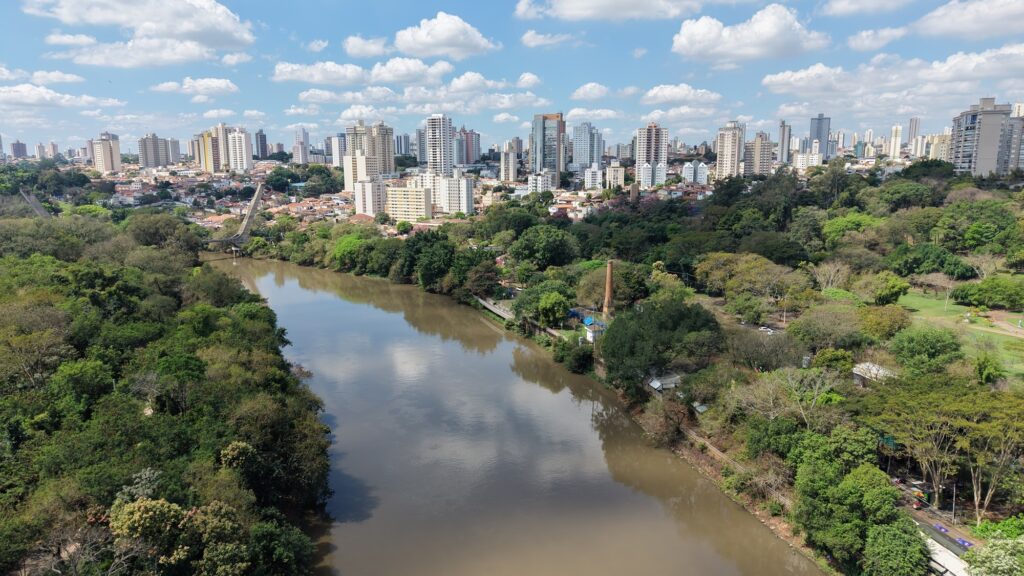 A imagem mostra uma vista panorâmica de uma cidade, com um rio de águas barrentas atravessando uma área bastante arborizada. Às margens, há parques, trilhas, uma antiga chaminé de tijolos e uma pequena ponte. Ao fundo, aparecem muitos prédios altos misturados a casas de telhados vermelhos. O céu está azul, com várias nuvens brancas, criando uma paisagem ensolarada que combina natureza e área urbana.