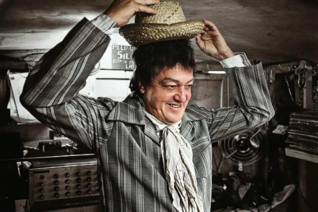 Um homem de meia-idade sorri enquanto coloca um chapéu de palha na cabeça em um ambiente interno bagunçado. A imagem retrata um homem de meia-idade com cabelo escuro e ondulado e uma expressão facial genuína de alegria enquanto ele tenta equilibrar um chapéu de palha na cabeça. Ele está vestindo uma camisa de manga comprida com um padrão de riscas verticais em tons de cinza e branco, com uma gola aberta. Um lenço de cor clara, amarrado solto no pescoço, adiciona um toque de estilo à sua vestimenta. Seus braços estão levantados enquanto ele ajeita o chapéu, com as mãos envolvendo a aba. O foco principal está no rosto do homem e no ato de colocar o chapéu. O ambiente ao redor parece ser um espaço interno desordenado, com vários objetos espalhados. À esquerda, há um aparelho eletrônico com botões e mostradores, sugerindo um ambiente como uma oficina ou um espaço de trabalho antigo. Atrás dele, à direita, pode ser visto um ventilador empoeirado e pilhas de objetos, possivelmente discos ou livros. Há também uma placa com texto parcialmente visível na parede ao fundo, escrita em português. A iluminação é um pouco dramática, com sombras acentuadas, o que contribui para uma atmosfera nostálgica e ligeiramente rústica. As cores predominantes são tons de cinza, branco e marrom, com o chapéu de palha adicionando um toque de cor terrosa. A composição, com o homem centrado e o fundo um pouco desfocado, realça o sujeito e sua ação. O humor geral da imagem é leve, divertido e caloroso, capturado pelo sorriso contagiante do homem.
