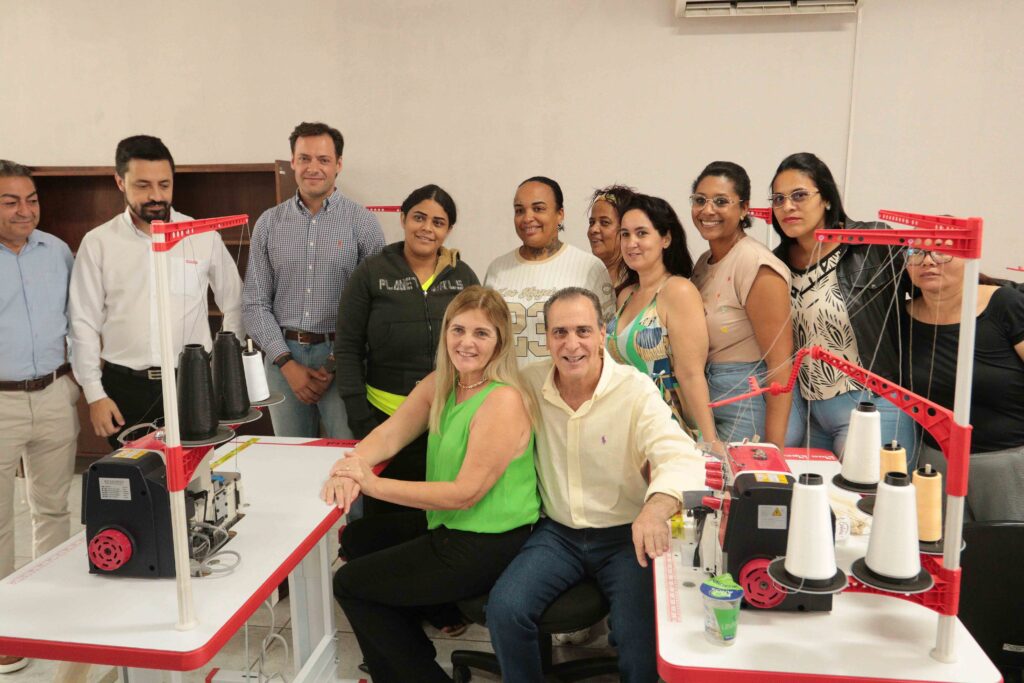 A imagem mostra uma oficina de costura ou bordado em grupo. Várias pessoas estão reunidas ao redor de mesas brancas, usando máquinas de costura e bordado industriais, principalmente nas cores branca, preta e vermelha. Há muitos carretéis de linha, tecidos e materiais de trabalho espalhados pelas mesas. Os participantes parecem concentrados na atividade; alguns usam camisetas com estampas coloridas e outros vestem aventais ou roupas casuais. O ambiente é interno, bem iluminado e organizado para uma aula ou treinamento prático.