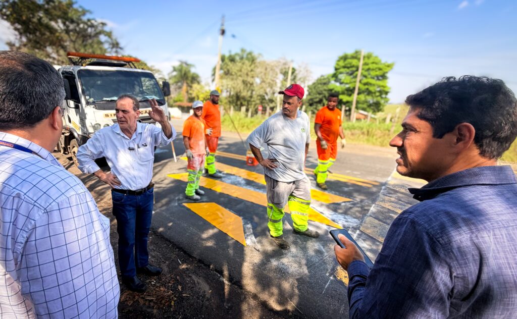A imagem mostra uma cena de trabalho em uma via pública, com vários homens reunidos ao redor de um caminhão de serviço. No centro, um homem de camisa azul-clara parece conversar com outras pessoas. À direita, há trabalhadores usando uniformes laranja com faixas refletivas, próximos a uma faixa de pedestres recém-pintada de amarelo. Em primeiro plano, aparecem partes de outras pessoas e um grande equipamento ou veículo escuro. O local tem árvores, postes e céu azul ao fundo, sob forte luz do dia.