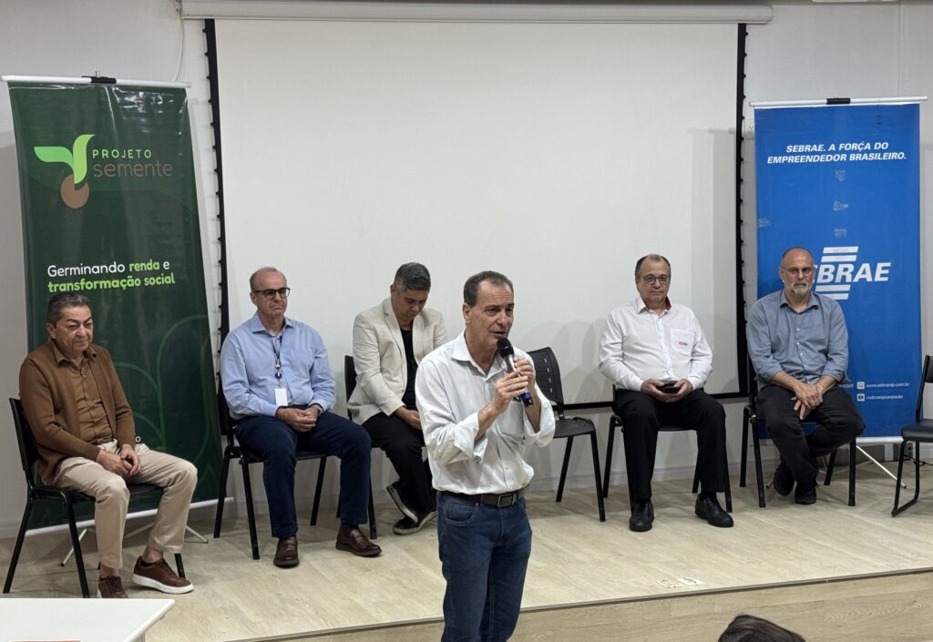 A imagem mostra um evento ou palestra em uma sala. Em primeiro plano, um homem de camisa branca e calça jeans segura um microfone e fala com o público. Atrás dele, há cinco homens sentados em cadeiras, aparentemente participando de um painel. Ao fundo, aparecem dois banners: um verde do Projeto Semente, com a frase “Germinando renda e transformação social”, e outro azul do Sebrae, com o texto “A força do empreendedor brasileiro”.