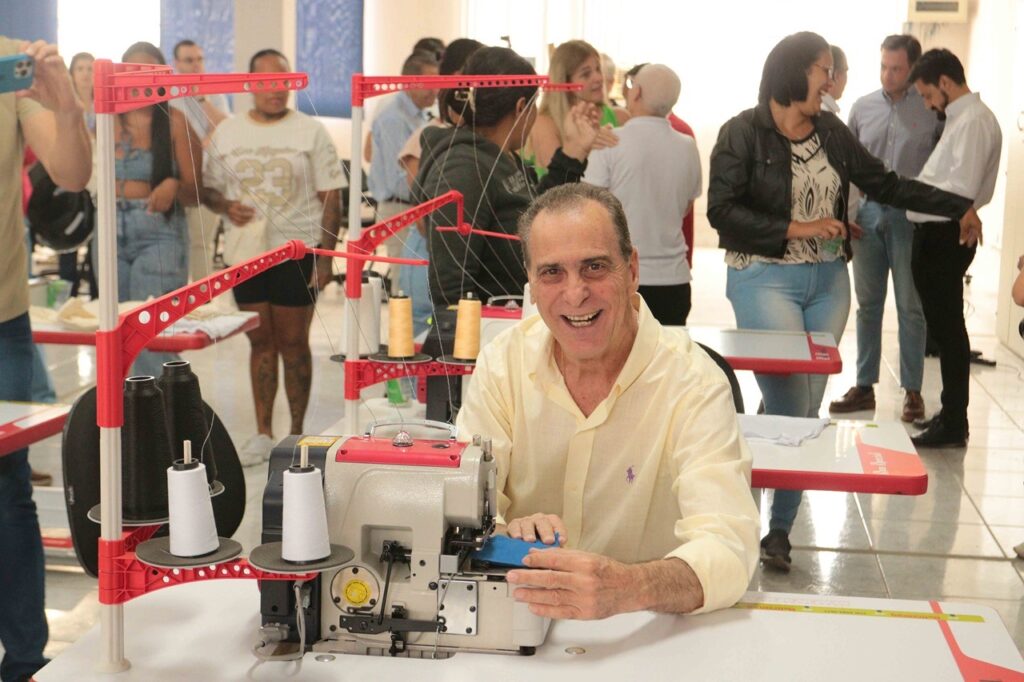 A imagem mostra um homem sorrindo enquanto trabalha em uma máquina de costura industrial. Ele está sentado diante de uma mesa branca, com as mãos próximas ao tecido e à área de costura. Ao redor da máquina há vários carretéis de linha preta, branca e bege, organizados em suportes vermelhos. Ao fundo, várias pessoas conversam e observam a atividade em um ambiente amplo e bem iluminado, provavelmente durante uma oficina ou evento de costura.