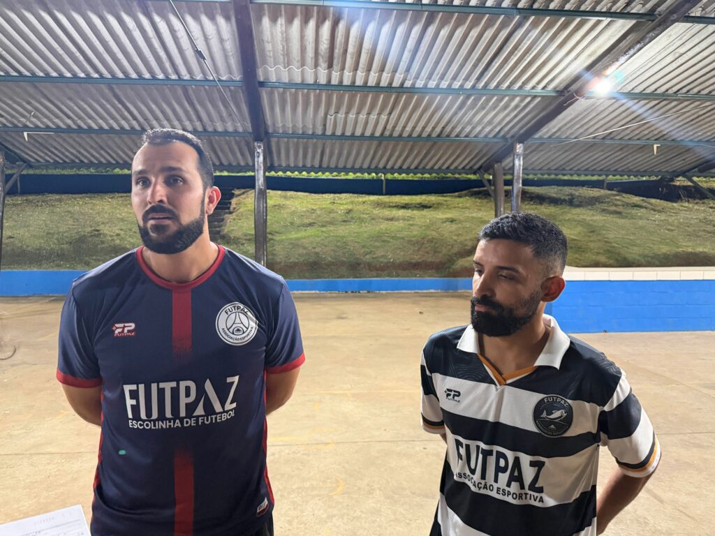 Duas pessoas usando camisetas de uma associação esportiva posam para a foto em um espaço coberto, durante o evento comunitário.