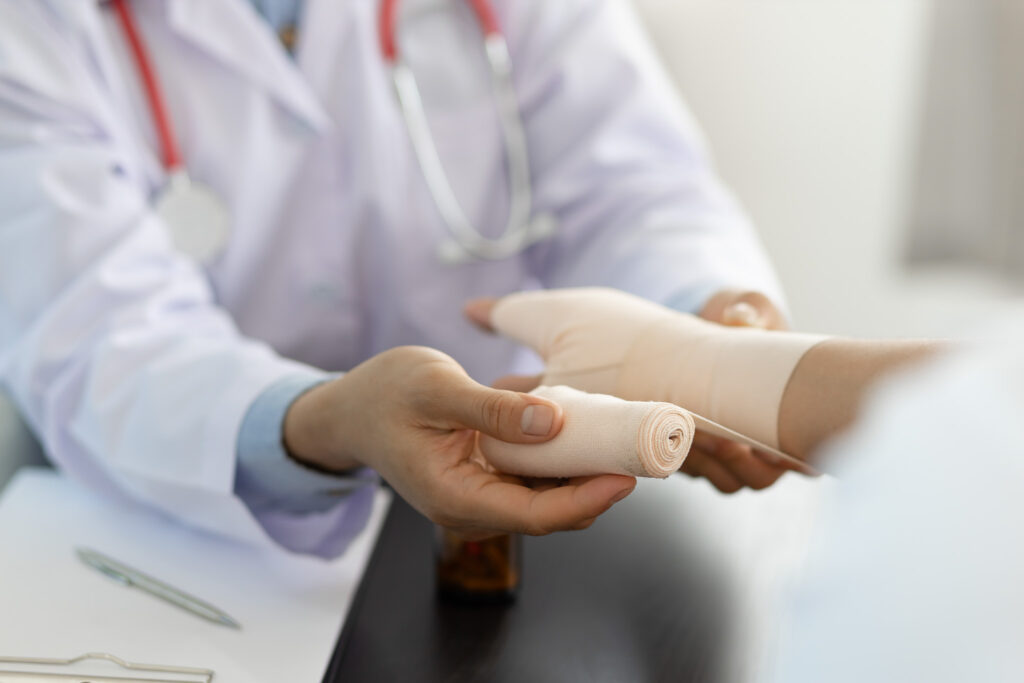 Profissional da área da saúde aplicando ou preparando uma bandagem na mão ou no punho de uma pessoa em um ambiente clínico. A cena mostra um jaleco, um estetoscópio e materiais médicos ao fundo.
