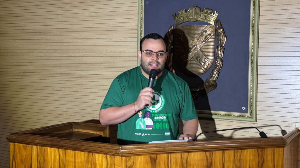 A imagem mostra um palestrante em um púlpito de madeira, segurando um microfone e falando para o público. Ele veste uma camiseta verde com o logotipo da CIPA (Comissão Interna de Prevenção de Acidentes) e os dizeres “prevenção, saúde e bem-estar”, indicando que o evento está relacionado à segurança do trabalho ou à promoção da saúde ocupacional.