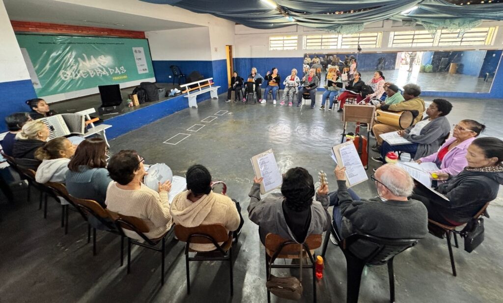 A imagem mostra um grupo de pessoas reunidas em uma sala ampla, sentadas em círculo durante uma atividade musical. Algumas seguram folhas com letras ou partituras; outras tocam instrumentos, como acordeão, pandeiro, chocalhos e tambores. As paredes são pintadas de azul e branco, e há um grande espelho ao fundo. No palco, aparece uma projeção com o texto “Oficinas de Bandas – Piracicaba”. O teto está decorado com tecidos escuros e pequenas luzes.
