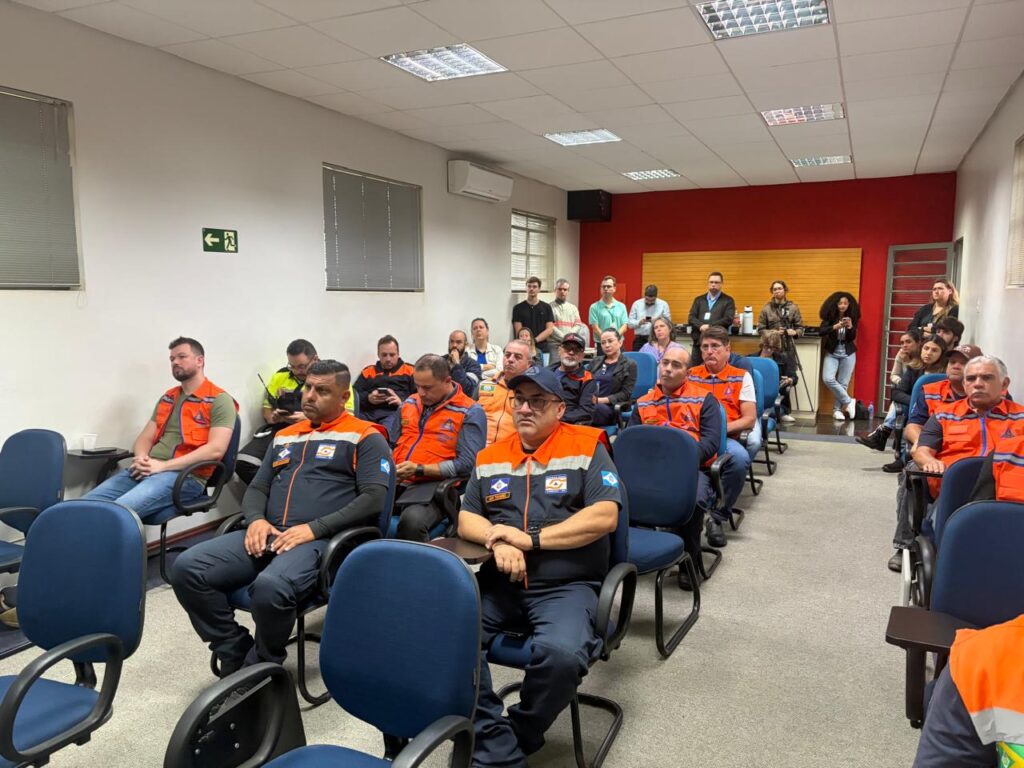 A imagem mostra uma sala de reunião ou treinamento com várias pessoas sentadas e outras em pé ao fundo. A maioria veste uniformes da Defesa Civil, com coletes laranja e detalhes em azul, enquanto alguns participantes usam roupas casuais. O ambiente é bem iluminado, com paredes brancas e uma parede vermelha ao fundo, onde há uma bancada com garrafas térmicas e copos. O clima parece de atenção e aprendizado, indicando uma capacitação ou reunião técnica voltada à segurança e gestão de emergências.