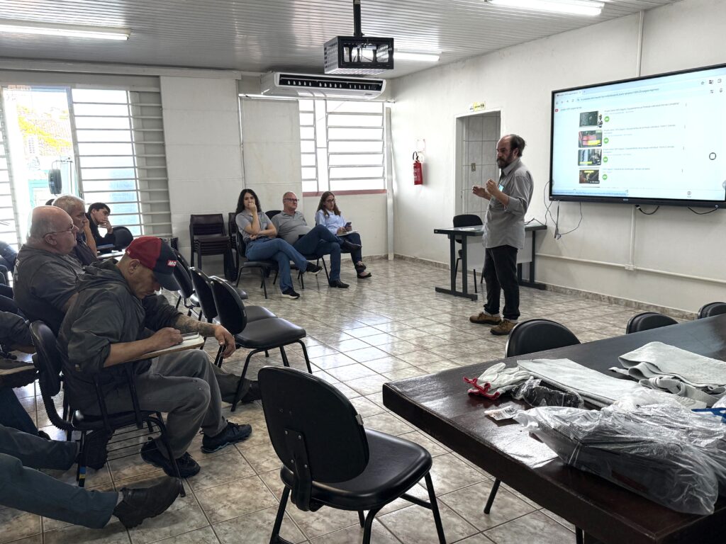 A imagem mostra uma aula ou treinamento em andamento. Um homem está em pé, falando para um pequeno grupo de pessoas sentadas em cadeiras, algumas tomando notas e outras observando atentamente. No fundo, há uma tela digital exibindo vídeos, sugerindo o uso de recursos audiovisuais na apresentação. O ambiente é simples, com piso de cerâmica, paredes brancas e iluminação fluorescente — típico de uma sala de reuniões ou espaço de capacitação profissional.