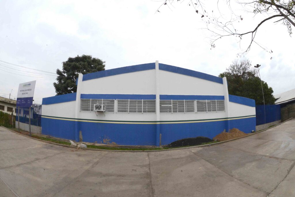 A imagem mostra um edifício público com uma pintura azul e branca, com uma faixa verde e amarela no meio, sugerindo que a construção está em reforma. A imagem exibe um edifício de um só piso com uma fachada imponente pintada em azul vibrante na parte inferior e branca na parte superior, separadas por uma fina linha horizontal verde e amarela. O edifício possui múltiplas janelas gradeadas, com um ar condicionado instalado em uma delas, indicando possivelmente um espaço comercial ou administrativo. À esquerda, um letreiro destaca o nome "PIRACICABA" e a frase "REFORMA DOS PRÉDIOS MUNICIPAIS", confirmando a natureza da obra. O local parece ser um pátio ou estacionamento asphaltado, com algumas áreas de terra e grama ao redor. Uma árvore com galhos finos e poucas folhas se projeta do canto superior direito, contra um céu nublado e claro. A iluminação é difusa, típica de um dia encoberto, criando uma atmosfera neutra e focada na estrutura do edifício e na informação do letreiro. Não há pessoas ou animais visíveis na cena. O humor geral da imagem é informativo e funcional, destacando um projeto de infraestrutura urbana.
