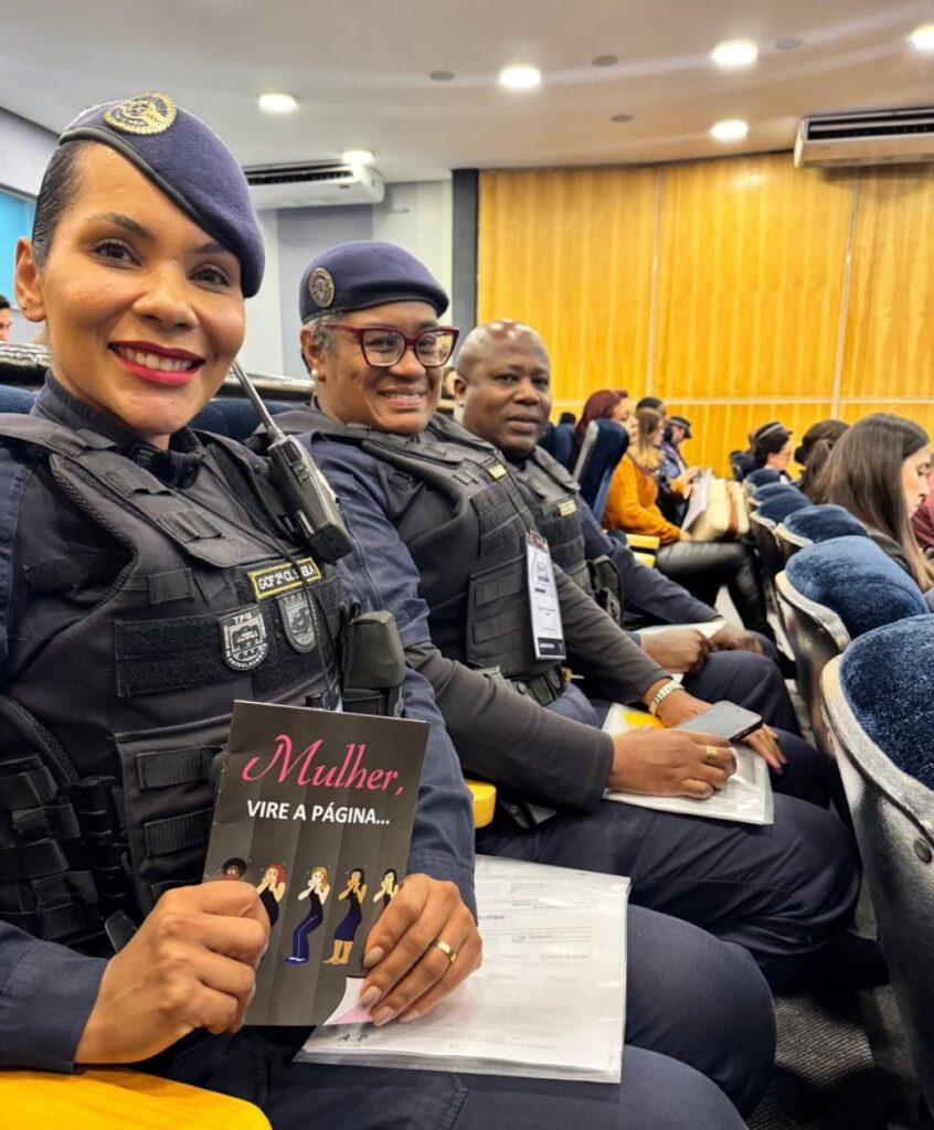 A imagem mostra três policiais fardados sentados em um auditório, sorrindo e participando de um evento. Eles vestem uniformes escuros com coletes táticos e boinas, indicando uma força de segurança. A policial em primeiro plano segura um livreto com o título “Mulher, vire a página...”, sugerindo que o encontro aborda temas de valorização feminina ou conscientização social.