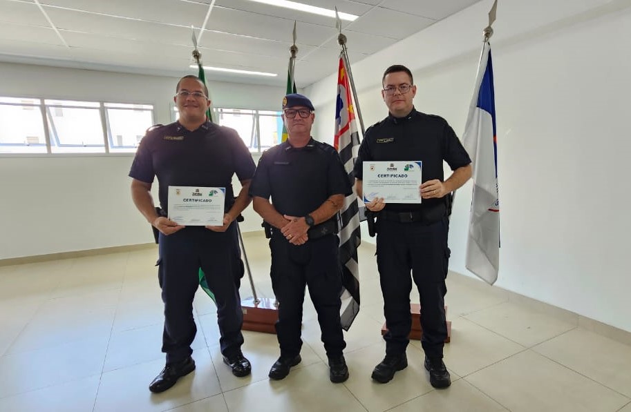 A imagem mostra três agentes uniformizados posando para uma foto em um ambiente institucional. Duas pessoas seguram certificados, enquanto a pessoa ao centro está posicionada diante de bandeiras oficiais. O registro sugere uma cerimônia de reconhecimento, capacitação ou entrega de certificados relacionada à área de segurança pública.